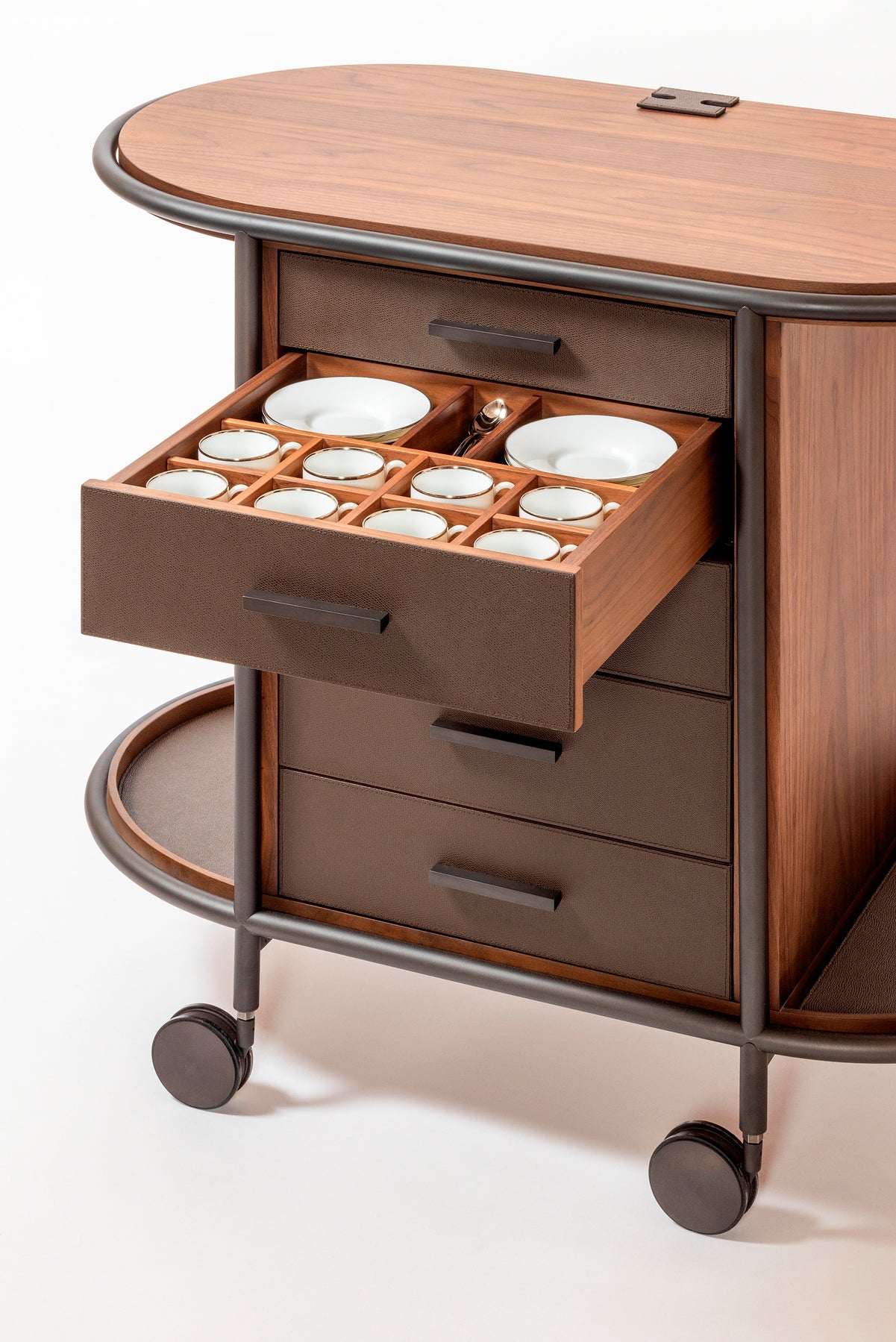 Giobagnara Arcade wood coffee and tea cart oval | Refined coffee and tea cart with a four-wheel structure in natural, walnut, or wenge wood | Metal frame and wheels available in chrome, bronze, or brass finishes | Features five organized drawers with sleek metal handles for storing coffee capsules, tea bags, and accessories | Includes porcelain coffee and tea cups with matching plates and silver graphic trim, plus wood trays and coasters | Designed for easy mobility and sophisticated presentation | Perfect 