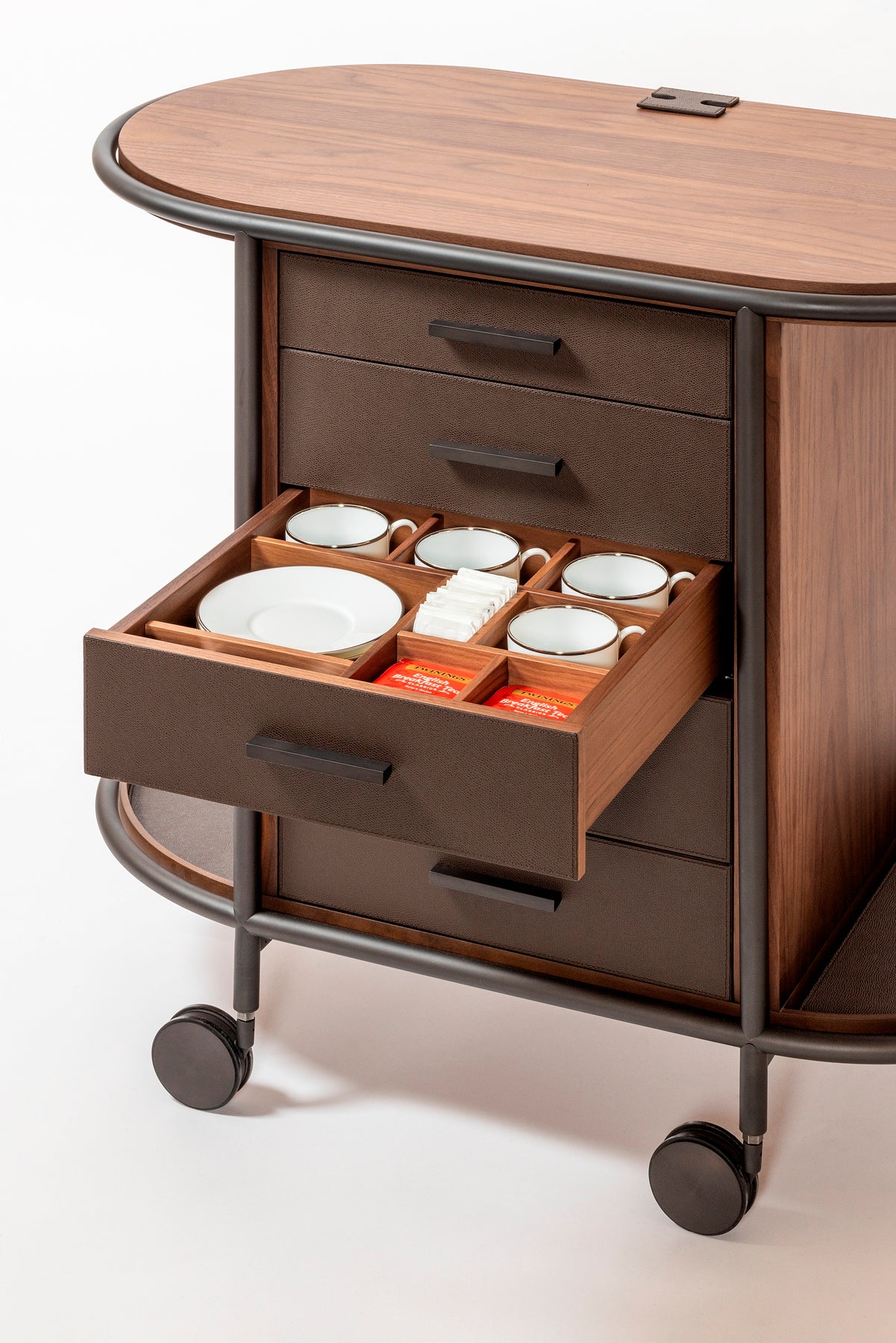 Giobagnara Arcade wood coffee and tea cart oval | Refined coffee and tea cart with a four-wheel structure in natural, walnut, or wenge wood | Metal frame and wheels available in chrome, bronze, or brass finishes | Features five organized drawers with sleek metal handles for storing coffee capsules, tea bags, and accessories | Includes porcelain coffee and tea cups with matching plates and silver graphic trim, plus wood trays and coasters | Designed for easy mobility and sophisticated presentation | Perfect 