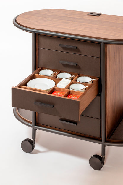 Giobagnara Arcade wood coffee and tea cart oval | Refined coffee and tea cart with a four-wheel structure in natural, walnut, or wenge wood | Metal frame and wheels available in chrome, bronze, or brass finishes | Features five organized drawers with sleek metal handles for storing coffee capsules, tea bags, and accessories | Includes porcelain coffee and tea cups with matching plates and silver graphic trim, plus wood trays and coasters | Designed for easy mobility and sophisticated presentation | Perfect 