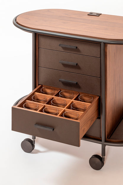 Giobagnara Arcade wood coffee and tea cart oval | Refined coffee and tea cart with a four-wheel structure in natural, walnut, or wenge wood | Metal frame and wheels available in chrome, bronze, or brass finishes | Features five organized drawers with sleek metal handles for storing coffee capsules, tea bags, and accessories | Includes porcelain coffee and tea cups with matching plates and silver graphic trim, plus wood trays and coasters | Designed for easy mobility and sophisticated presentation | Perfect 