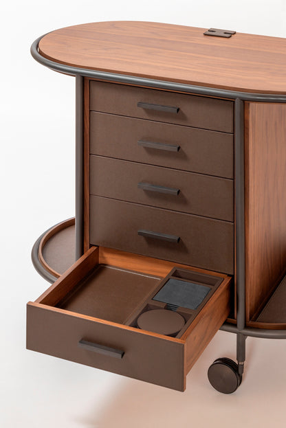 Giobagnara Arcade wood coffee and tea cart oval | Refined coffee and tea cart with a four-wheel structure in natural, walnut, or wenge wood | Metal frame and wheels available in chrome, bronze, or brass finishes | Features five organized drawers with sleek metal handles for storing coffee capsules, tea bags, and accessories | Includes porcelain coffee and tea cups with matching plates and silver graphic trim, plus wood trays and coasters | Designed for easy mobility and sophisticated presentation | Perfect 