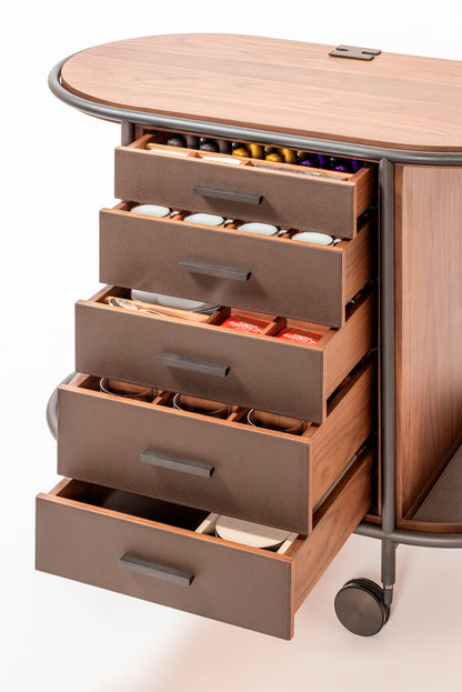 Giobagnara Arcade wood coffee and tea cart oval | Refined coffee and tea cart with a four-wheel structure in natural, walnut, or wenge wood | Metal frame and wheels available in chrome, bronze, or brass finishes | Features five organized drawers with sleek metal handles for storing coffee capsules, tea bags, and accessories | Includes porcelain coffee and tea cups with matching plates and silver graphic trim, plus wood trays and coasters | Designed for easy mobility and sophisticated presentation | Perfect 