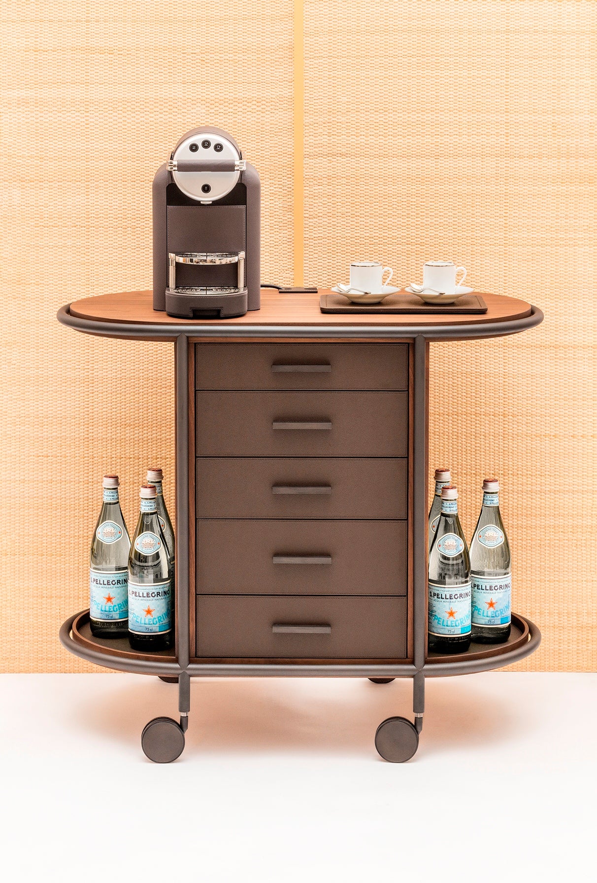 Giobagnara Arcade wood coffee and tea cart oval | Refined coffee and tea cart with a four-wheel structure in natural, walnut, or wenge wood | Metal frame and wheels available in chrome, bronze, or brass finishes | Features five organized drawers with sleek metal handles for storing coffee capsules, tea bags, and accessories | Includes porcelain coffee and tea cups with matching plates and silver graphic trim, plus wood trays and coasters | Designed for easy mobility and sophisticated presentation | Perfect 