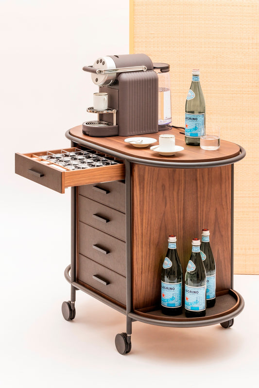 Giobagnara Arcade wood coffee and tea cart oval | Refined coffee and tea cart with a four-wheel structure in natural, walnut, or wenge wood | Metal frame and wheels available in chrome, bronze, or brass finishes | Features five organized drawers with sleek metal handles for storing coffee capsules, tea bags, and accessories | Includes porcelain coffee and tea cups with matching plates and silver graphic trim, plus wood trays and coasters | Designed for easy mobility and sophisticated presentation | Perfect 